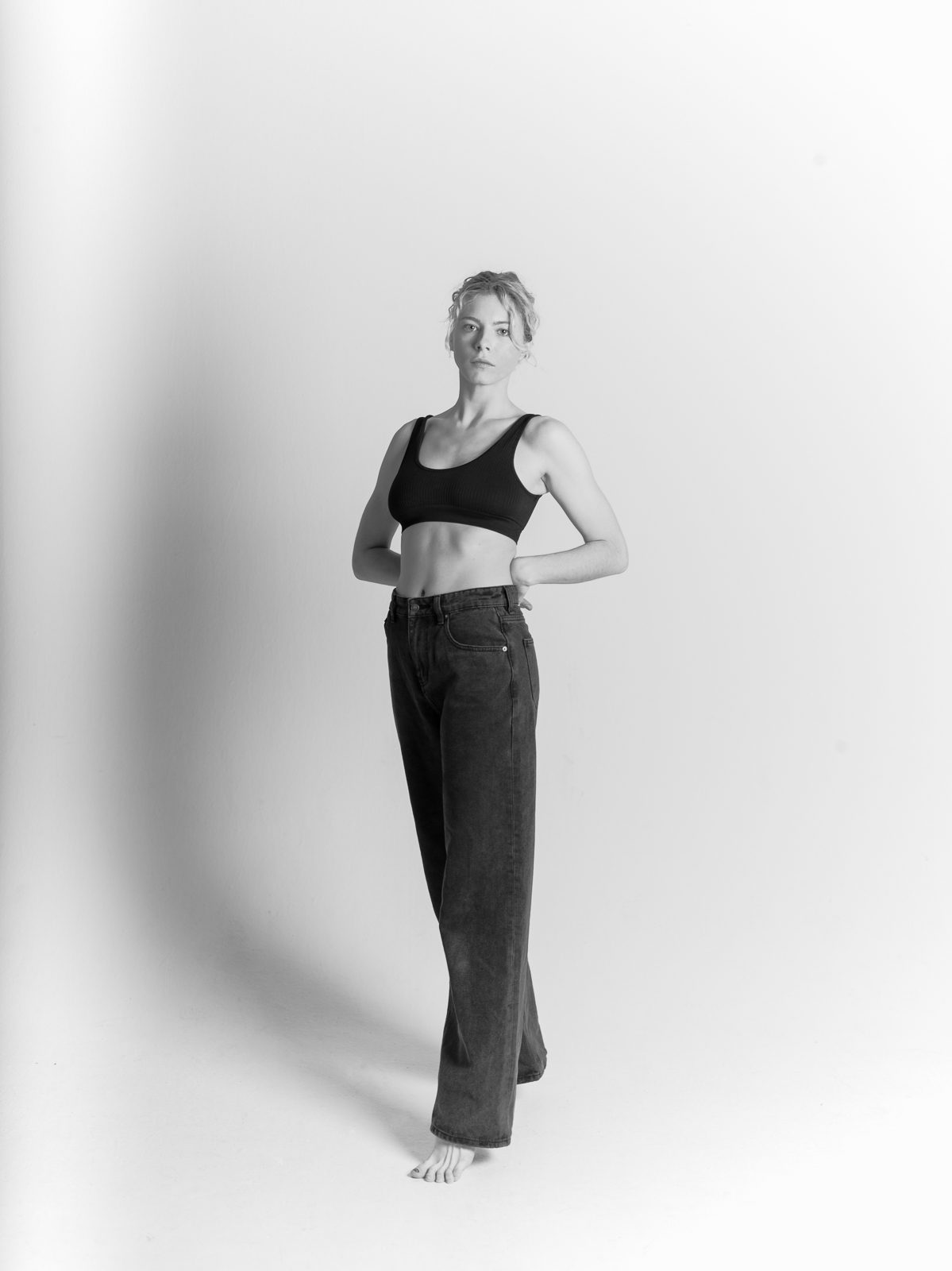 Frau im Studio in Bralette und weiter Jeans, Hand in der Tasche, heller Hintergrund, Schwarzweiß
